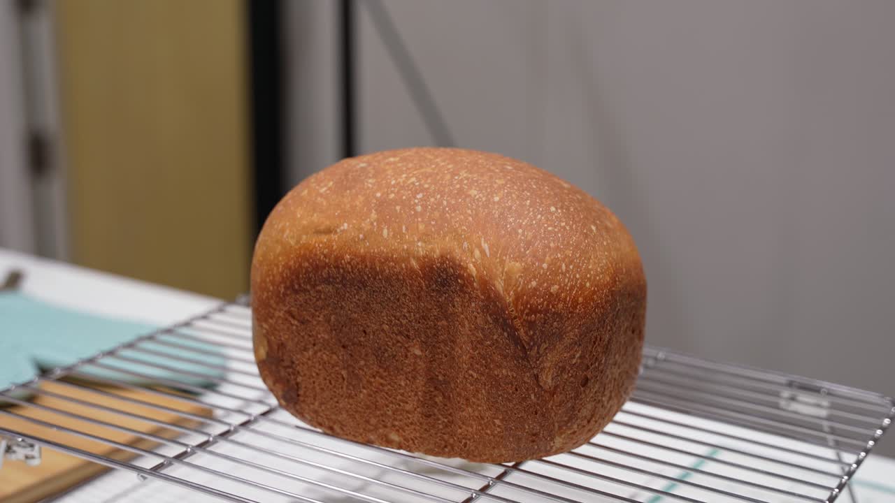 Close-up of a freshly baked bread loaf cooling on a wire rack indoors, showing golden brown crust, homemade baking results, and a clean kitchen background