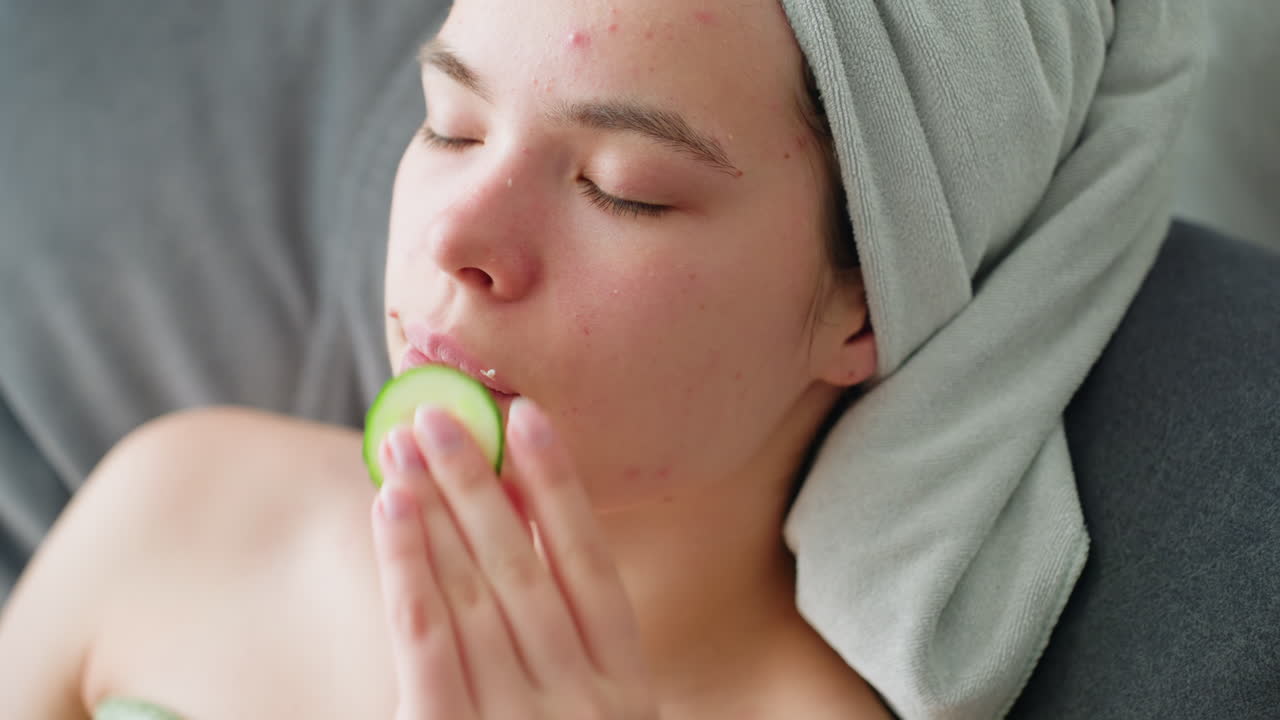Woman with towel-wrapped hair gently massaging face with cucumber slice as part of soothing skincare routine, captured in peaceful indoor setting focused on rejuvenation and natural beauty