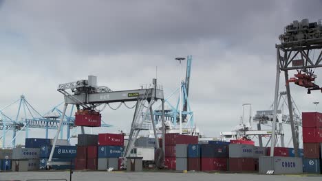 Vista-De-La-Terminal-De-Contenedores-De-Envío-Que-Muestra-Grúas-En-Azul-Y-Blanco,-Contenedores-Coloridos-En-Una-Disposición-Apilada-Sobre-Suelo-De-Concreto,-Bajo-Un-Cielo-Nublado