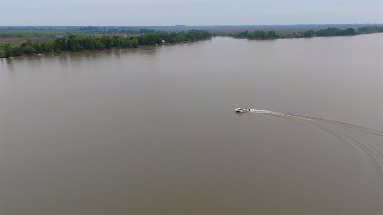 A white speedboat cuts through the wide muddy river in the Paraná Delta, leaving a curving wake behind as forest lines the far bank under a pale sky, aerial drone follow shot