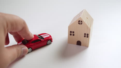 casa de juguetes de madera proyectando sombra en la mesa y luego los dedos izquierdos empujando en un coche deportivo de juguete rojo al lado de la casa de juguetes