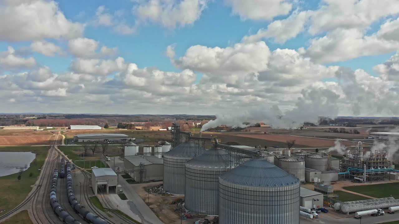 Aerial view of mill with steel silos for grain storage and processing plant, train tracks against beautiful rural countryside - drone ascending day