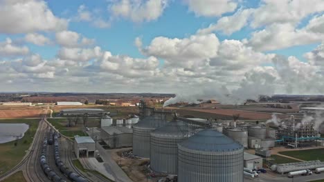 Aerial-view-of-mill-with-steel-silos-for-grain-storage-and-processing-plant,-train-tracks-against-beautiful-rural-countryside---drone-ascending-day