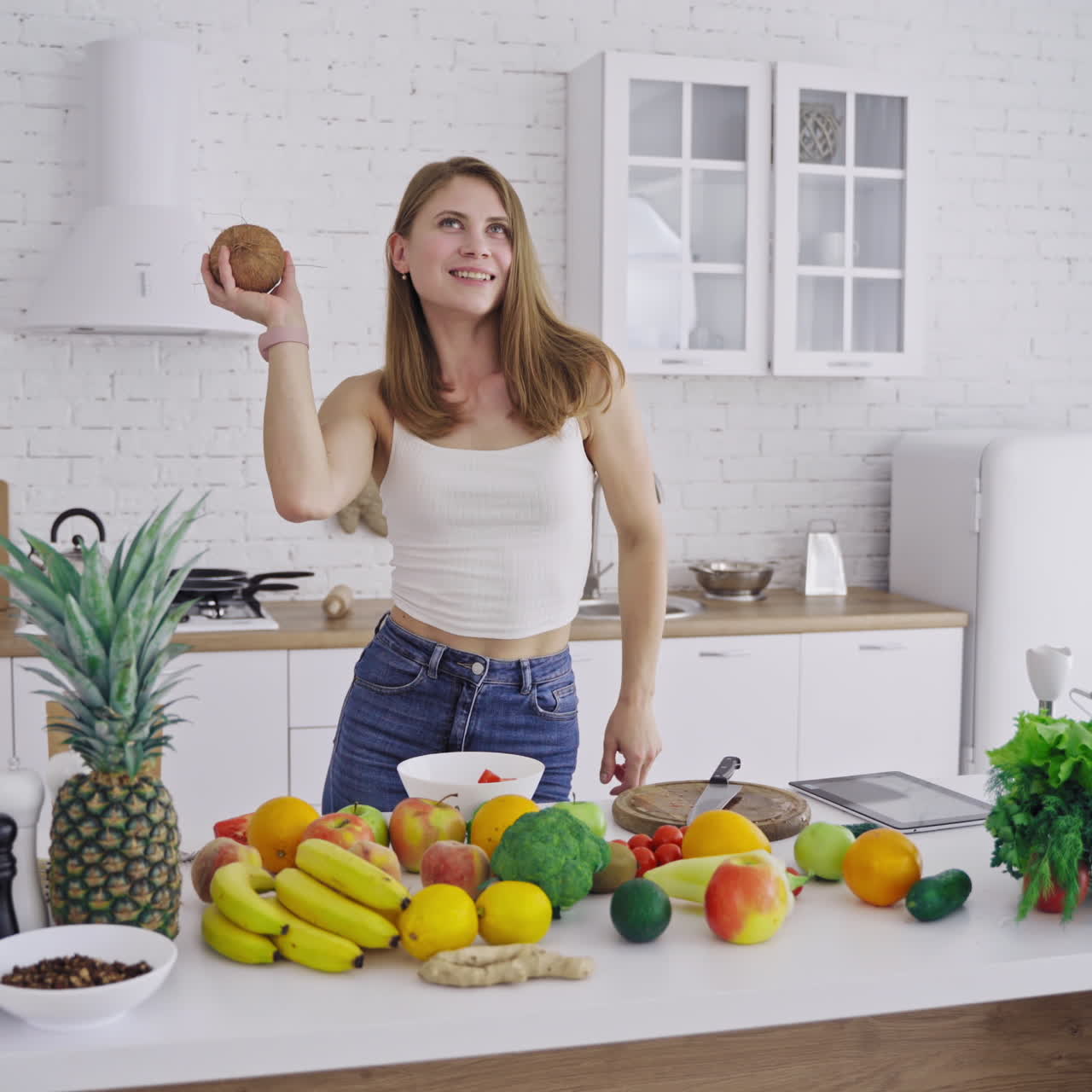 Joyful woman with coconut in the kitchen. Attractive female is dancing with fresh coconut on the background of organic fruit and vegetables. Healthy lifestyle.