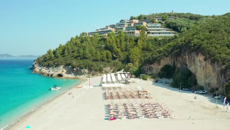 Drone-flyover-Karavostasi-Beach-towards-idyllic-Elix-Mar-Bella-Hotel-on-Forested-hillside,-Greece