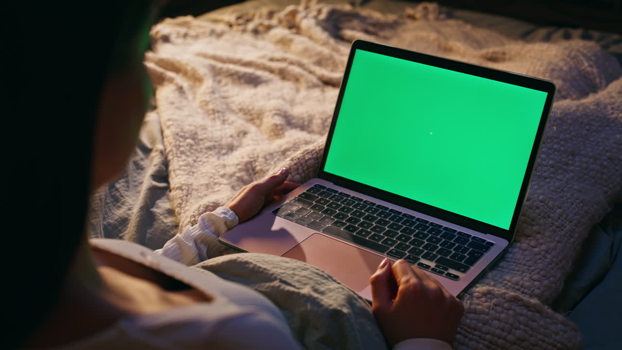 Primo piano di una freelance notturna che naviga su un laptop con schermo chroma key a letto. Donna che guarda il computer.