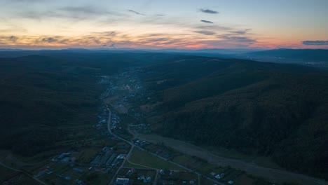 Toma-Aérea-De-Hiperlapso-De-Conducción-De-Automóviles-En-La-Carretera-Entre-El-Pueblo-En-Los-árboles-Del-Bosque-Al-Atardecer---Vuelo-Ascendente