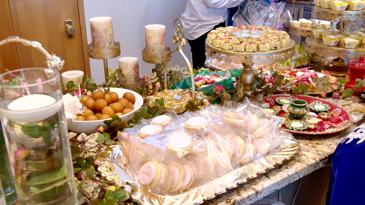 Ricco buffet di dolci con Gulab Jamun, pasticcini assortiti dell'Asia meridionale e dolcetti confezionati, impreziosito da portacandele dorati e decorati e centrotavola floreali galleggianti.