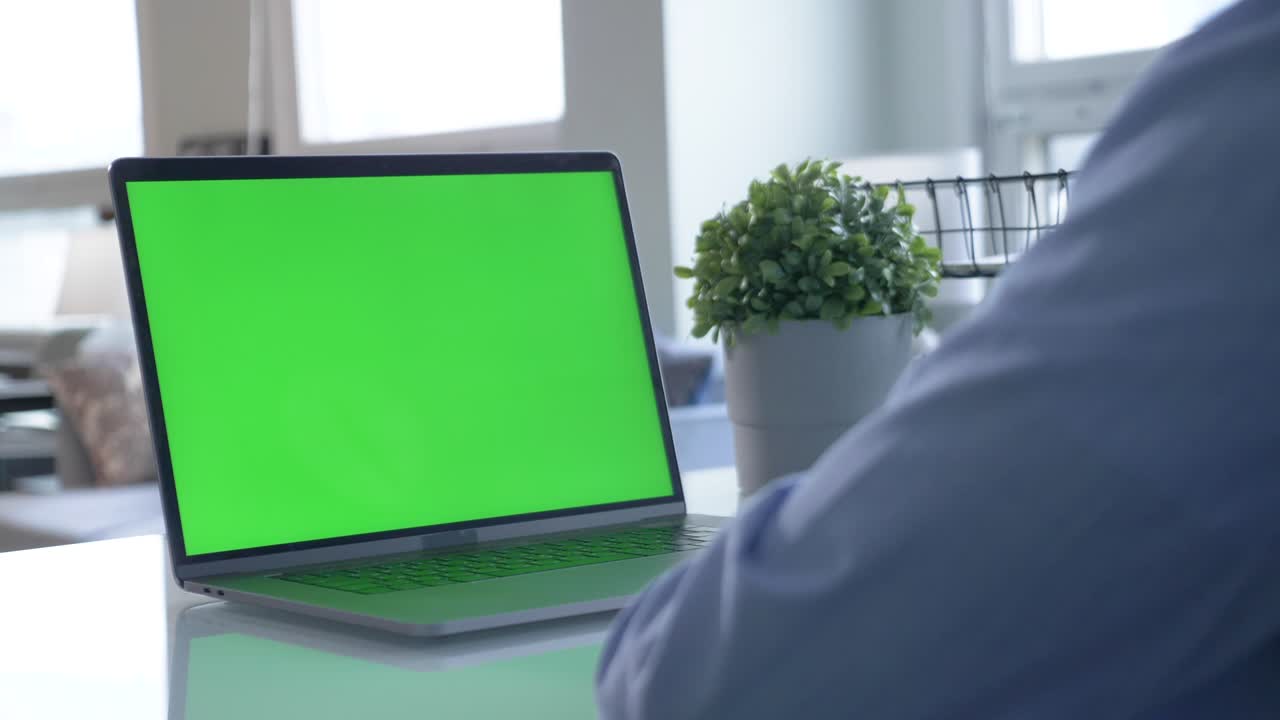 Male Office Worker at His Desk Works on a Laptop with Mock-up Green Screen. Over the Shoulder Close-up Footage. In the Background Creative Office