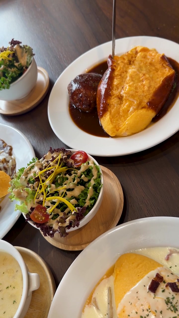Salsa ricca versata su riso all'omelette, hamburg steak, insalata, zuppa in un'ambientazione luminosa da caffè.