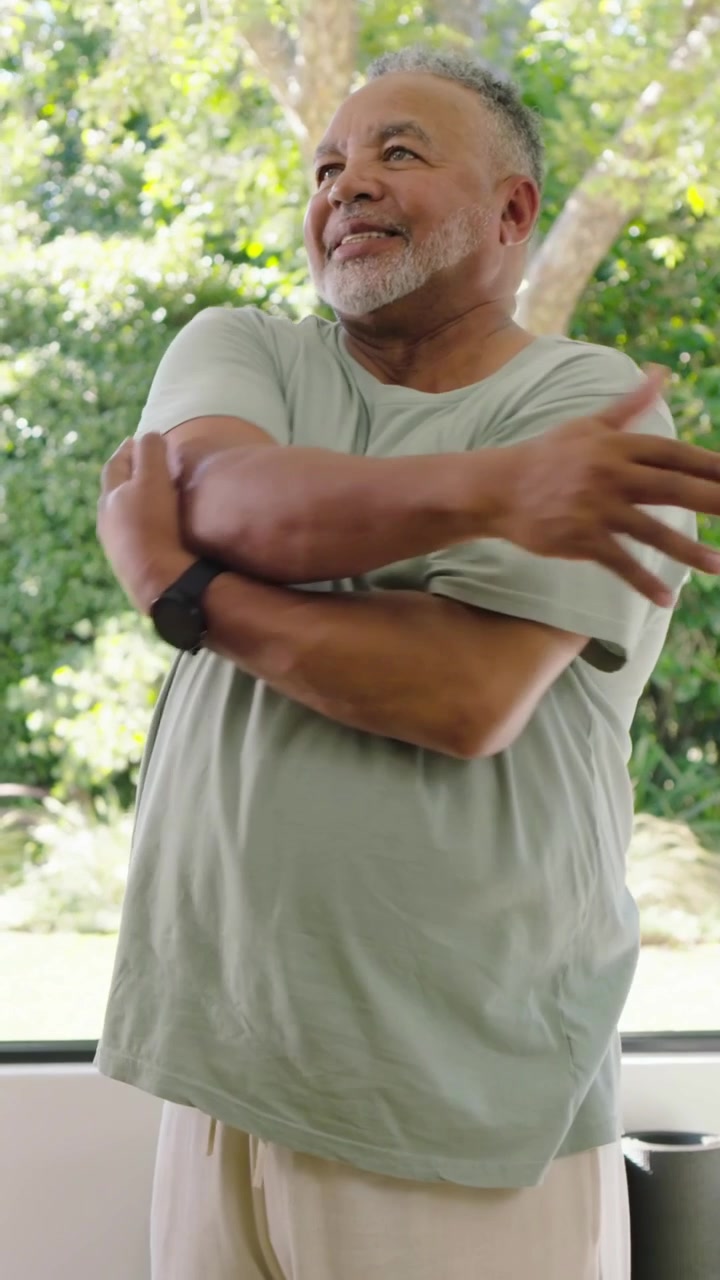 Vídeo vertical: Homem idoso se alongando com camiseta verde, cruzando o peito perto da janela, mão de assistente guiando.