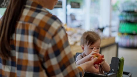 Nahaufnahme:-Eine-Mutter-In-Einem-Karierten-Hemd-Gibt-Ihrer-Kleinen-Tochter-Beim-Einkaufen-In-Einem-Supermarkt-Einen-Apfel.-Ein-Kleines-Mädchen-Hält-Einen-Apfel-In-Den-Händen-Und-Untersucht-Ihn-In-Einem-Supermarkt