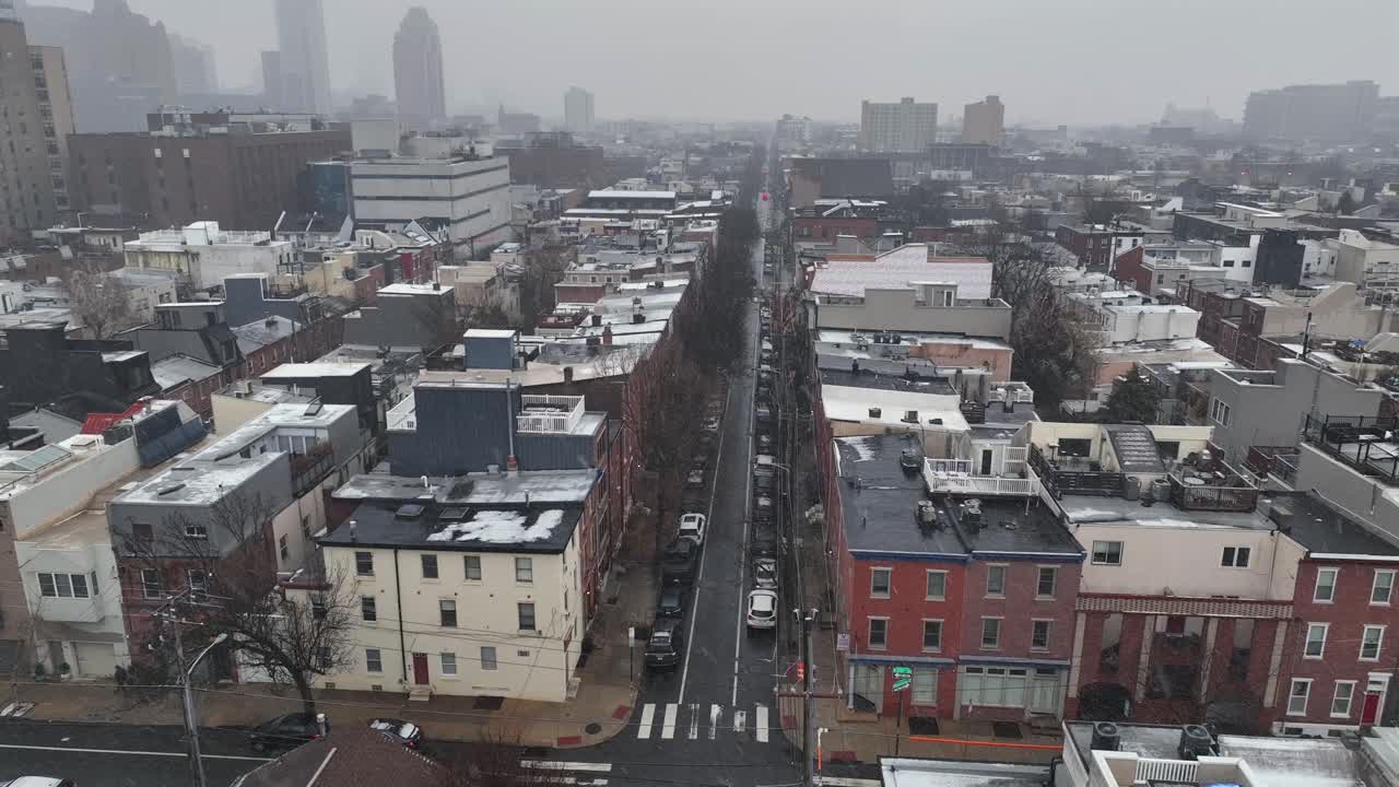 Luftfoto af et boligkvarter i Philadelphia om vinteren, der viser sneklædte tage, en lige bygade og en diset bysilhuet, der strækker sig ud i horisonten. Bredt billede