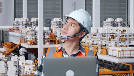 close up de ingeniero masculino asiático con casco de seguridad mirando una computadora portátil y mirando a su alrededor mientras está de pie en el almacén con estantes llenos de bienes de entrega