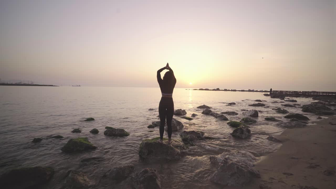 Giovane donna afroamericana atletica che pratica yoga all'alba su una roccia nell'oceano, routine di allenamento meditativo mattutino.
