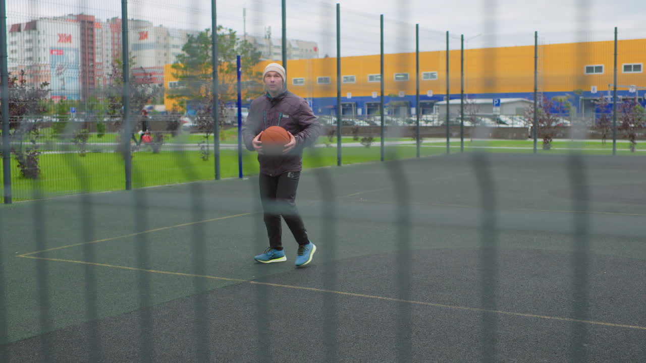 Un giocatore di basket urbano all'aperto fa una schiacciata, un dilettante solitario palleggia e scatta verso il canestro sotto un cielo nuvoloso in un parco giochi recintato con superficie asfaltata ed edifici commerciali gialli sullo sfondo