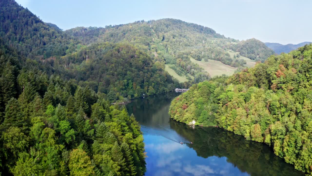 Dam at deep blue reflecting lake in hilly forest landscape, smooth aerial drone