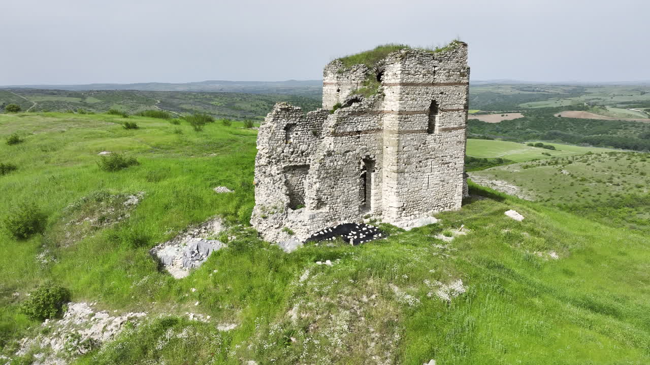 Orbiting drone view of a solitary medieval hilltop fortress surrounded by lush spring grass and rolling fields under a softly overcast sky