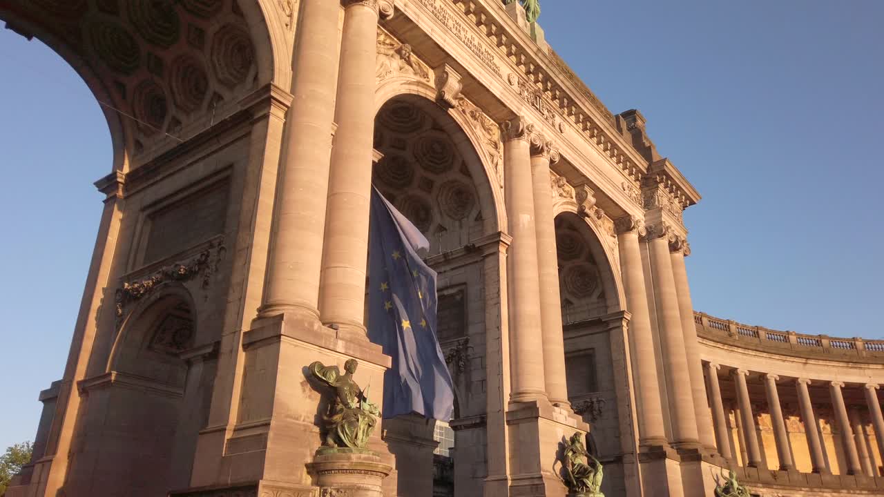 Wide-angle shot of the European Union flag waving beneath the iconic arches of the Cinquantenaire monument in Brussels, Belgium. The neoclassical structure is bathed in warm evening light