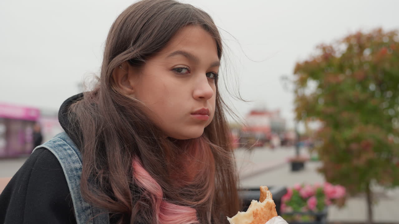Studente adolescente seduto su una panchina in un centro commerciale all'aperto che mangia un dolce con espressioni vivaci tra un morso e l'altro, sciarpa colorata e zaino di jeans, primi piani spontanei che mostrano curiosità e appetito giovanile sullo sfondo vivace