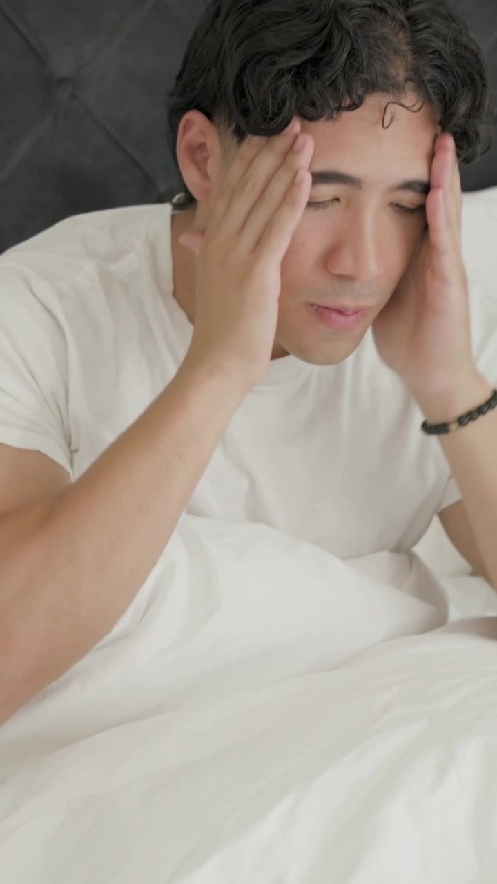Video vertikal: Pria terbangun dari tidur mengenakan kaus putih, menggosok mata dan duduk di tempat tidur, mencari istirahat.