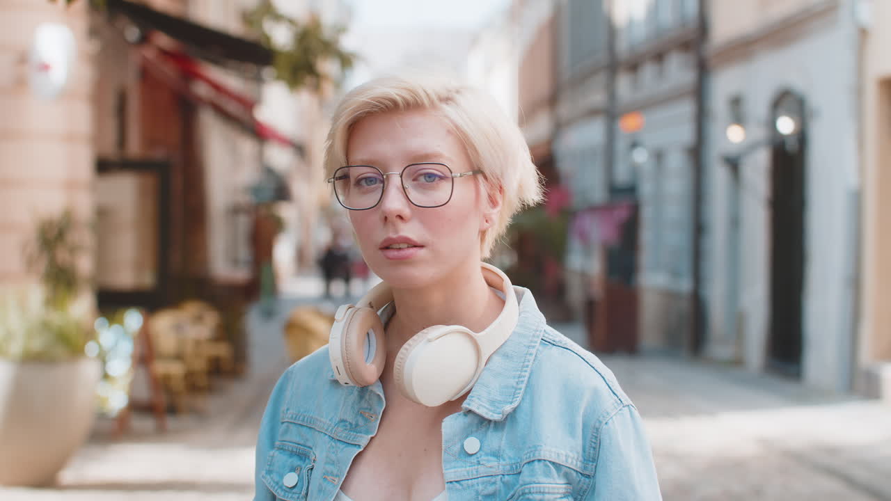 Portrait of smiling young woman in glasses taking off wireless headphones while looking at camera