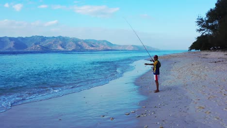 Young-man-fishing-a-small-fish-rolling-rod-line-from-blue-sea-on-empty-beach-at-sunset-low-light,-Bali