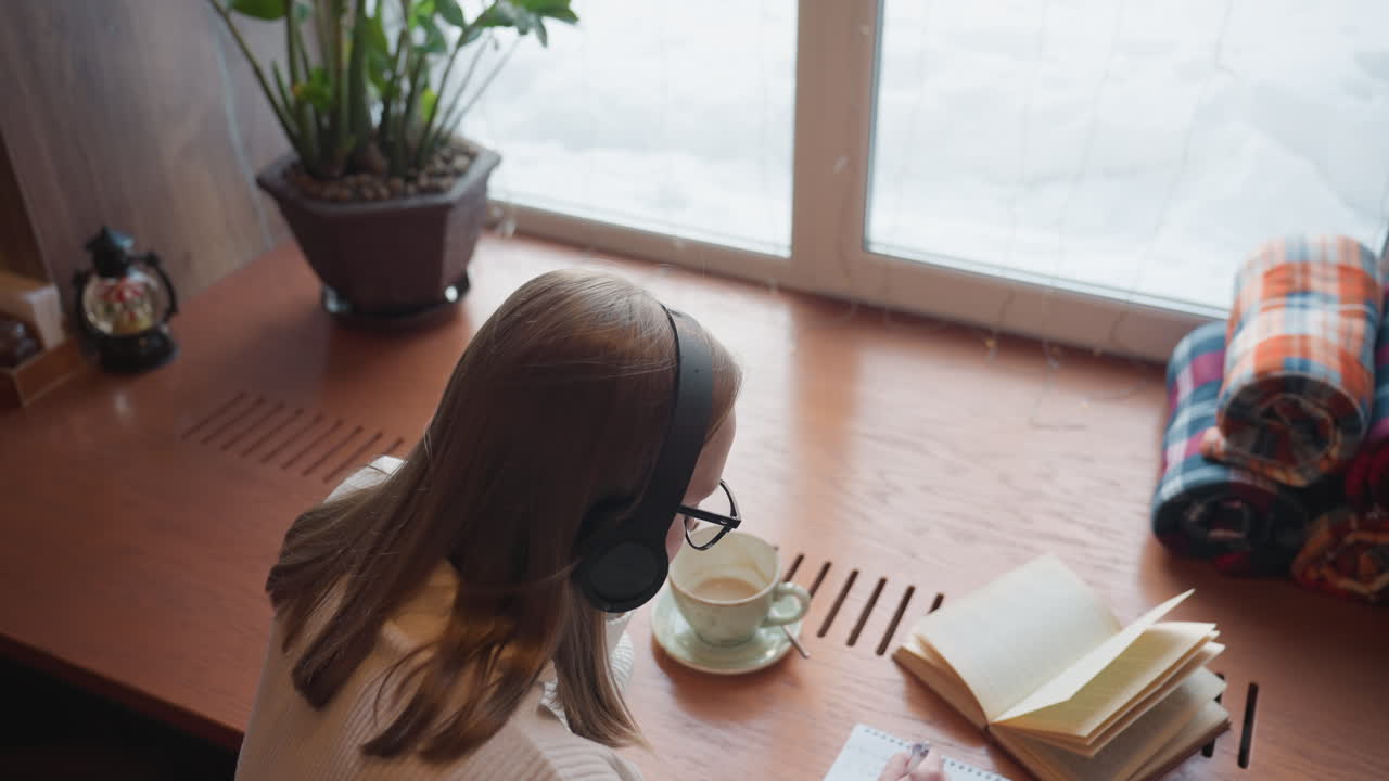 top down view of young lady in sweater wearing headphone studying and writing from open book while rhythmically moving to music beside mug and folded cloth on wooden table near window