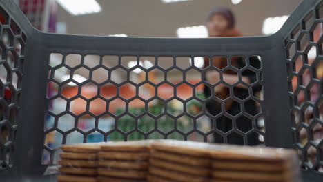 view of a lady in a black coat wearing a backpack placing a juice carton into a shopping trolley that already contains a pack of biscuits. capturing the shopping experience from in a grocery store