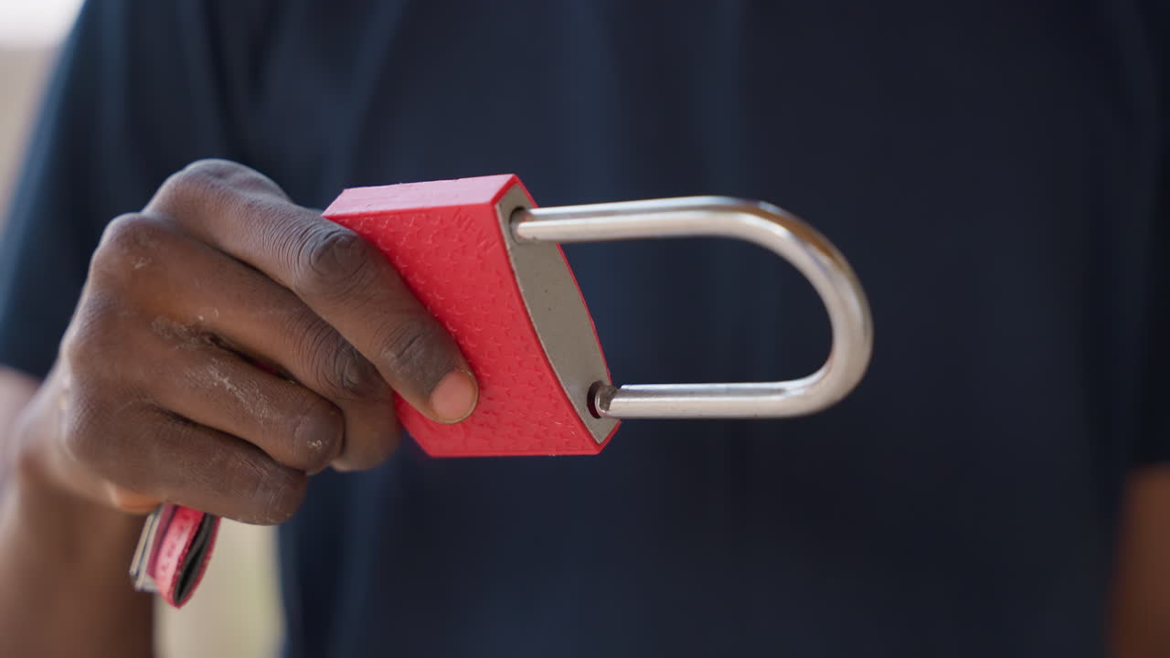 Closeup black man holding padlock, hands inspect shackle and textured red body while testing key and locking mechanism. Frames show careful grip, close examination, presentation to camera, and final
