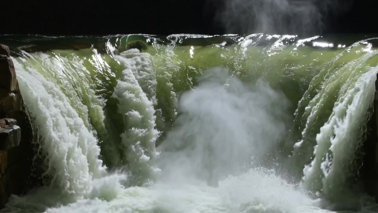 Una cascata fluida si getta dal bordo di notte, formando una colonna di nebbia che sale da un bacino spumeggiante, con una scogliera a sinistra.