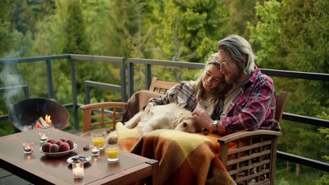 un hombre rubio con gafas y una chica rubia están sentados en un sofá y acariciando a su gran perro de color claro en el balcón de una casa de campo con vistas al bosque y las montañas