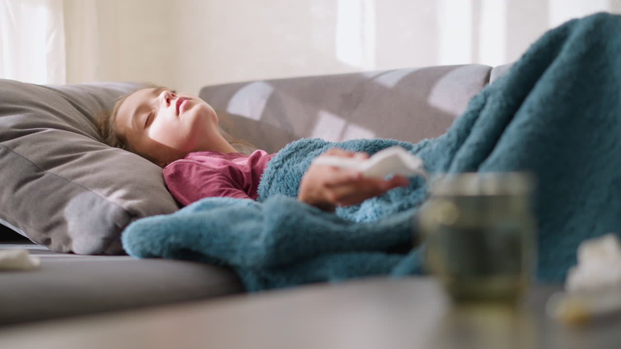 Bambina che si sistema sul divano prima di starnutire in un fazzoletto. Avvolta in una coperta, si sente male a casa, con fazzoletti sparsi intorno e una tazza appoggiata sul tavolo vicino