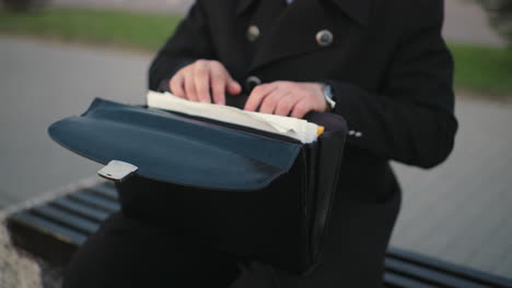 close up of individual hand in black clothing going through documents in bag, with background featuring greenery and interlocked path