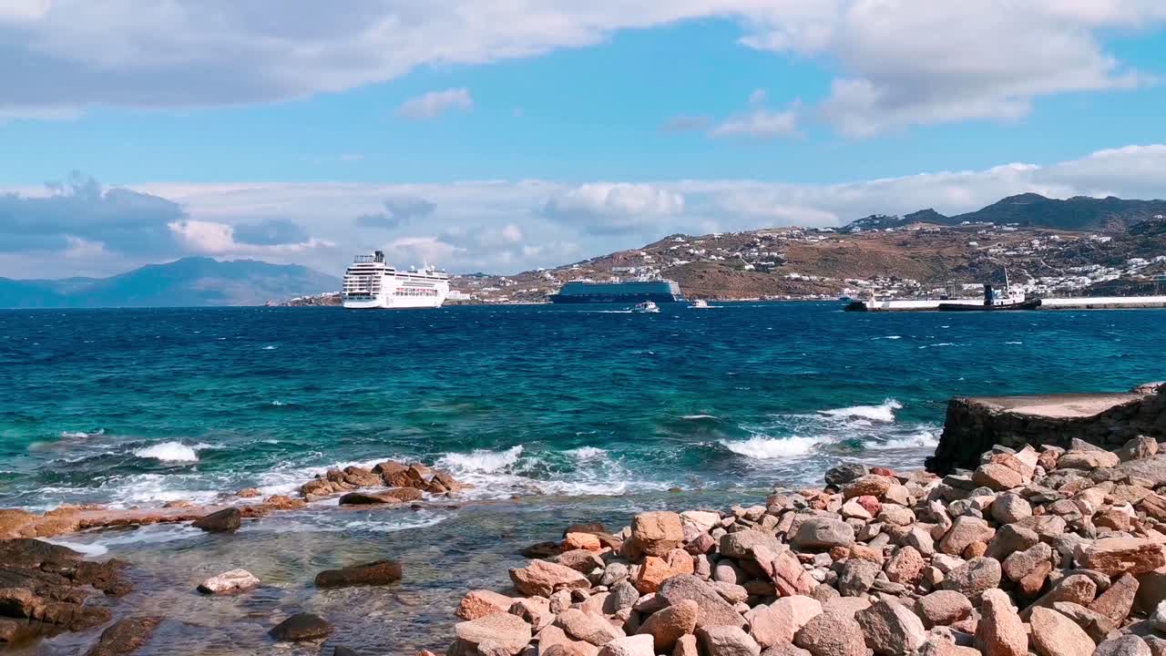 View of rocky beach in Santorini, Greece, with turquoise sea waves hitting shore, cruise ships in distance, and scenic coastal hills beneath a vibrant blue sky with scattered clouds
