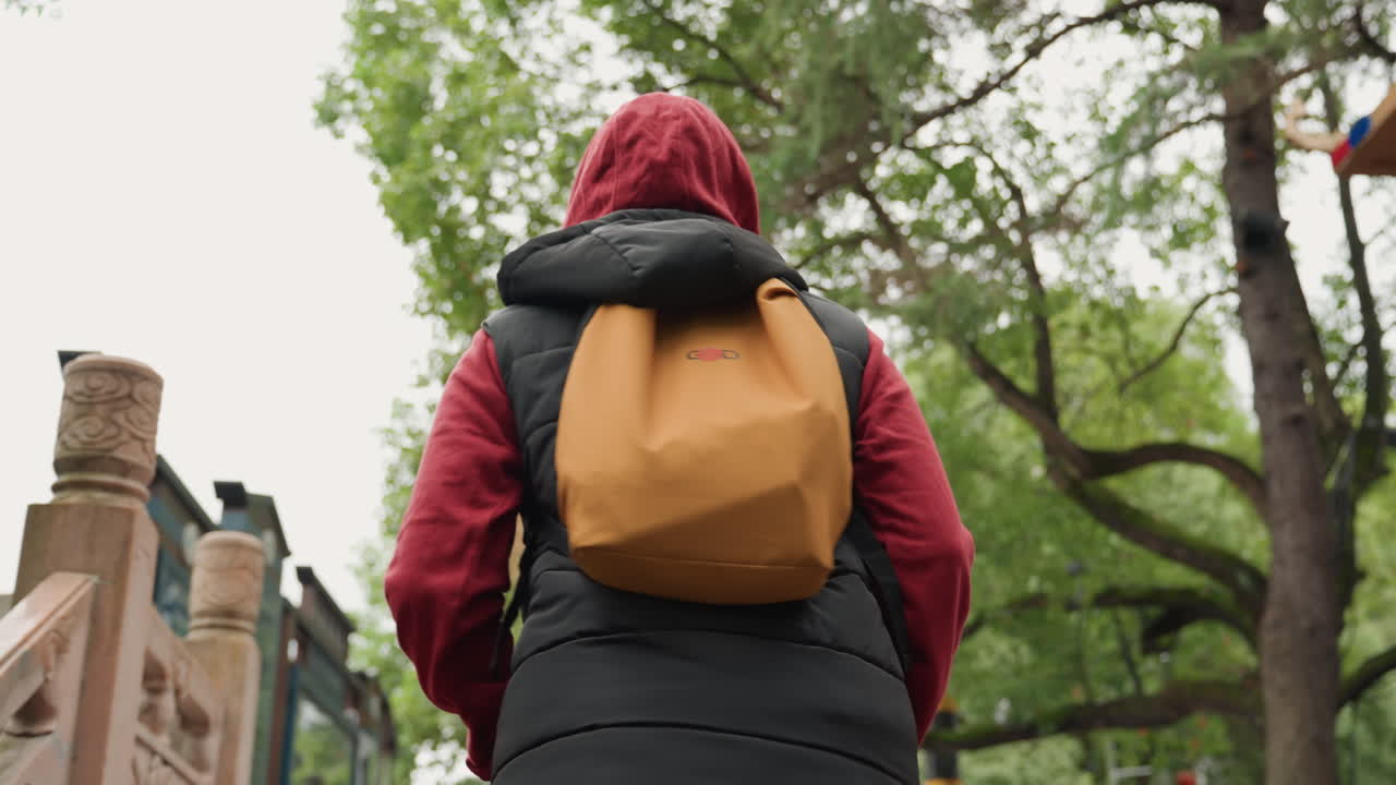City worker ascending. Individual with colorful backpack walking through lively urban area. Urban commuter with red jacket and yellow backpack captured from low angle in bustling city park