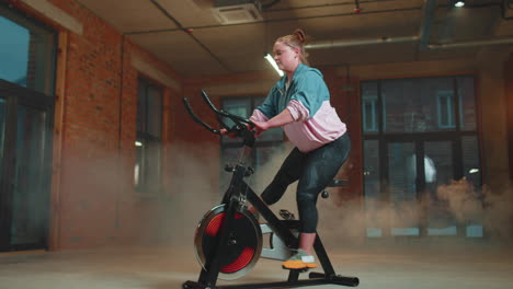 Athletic-girl-performing-aerobic-riding-training-exercises-on-cycling-stationary-bike-in-foggy-gym