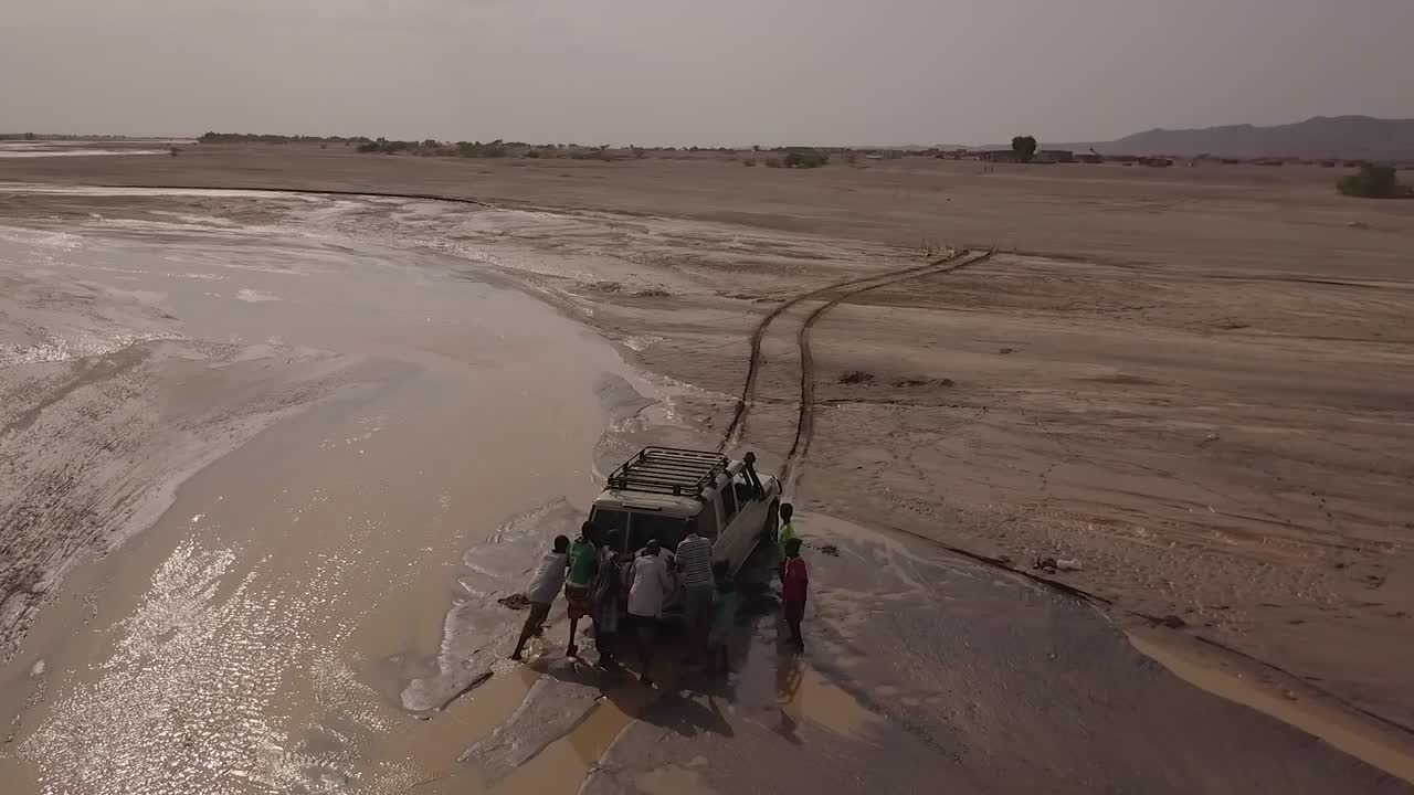 Ripresa aerea di persone che spingono una jeep 4x4 fuori da un fiume fangoso nei deserti di Gibuti o Somalia, Africa