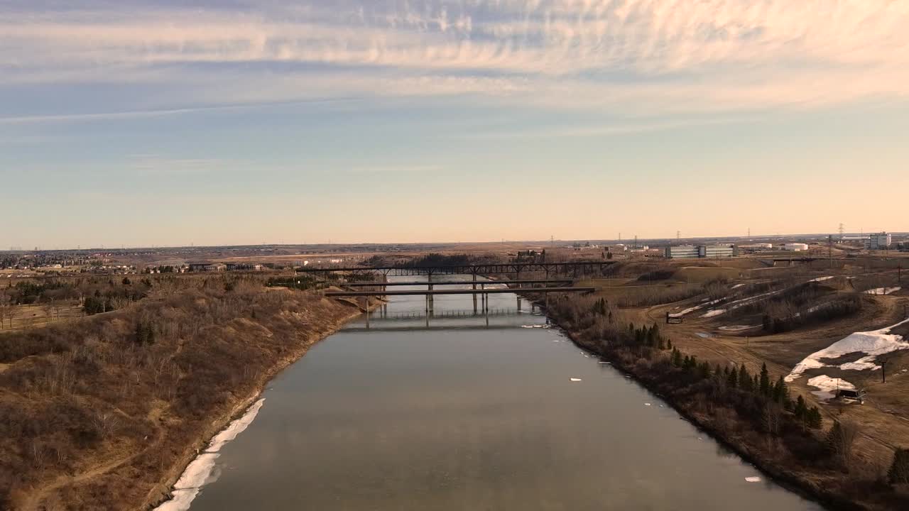 North Saskatchewan River flows toward Edmonton as early sunrise light casts warm tones across calm water and distant bridges creating peaceful prairie edge scene captured from steady drone view