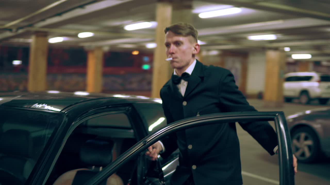 un joven con un traje negro y una pajarita fumando un cigarrillo, estacionando un coche y saliendo con una gran bolsa negra por la noche.