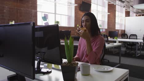 mixed race businesswoman sitting at desk eating pastries