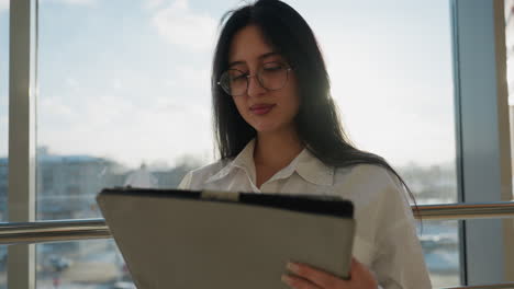 young woman in white shirt with glasses using tablet indoors near large glass window, natural daylight highlighting her calm expression with city background