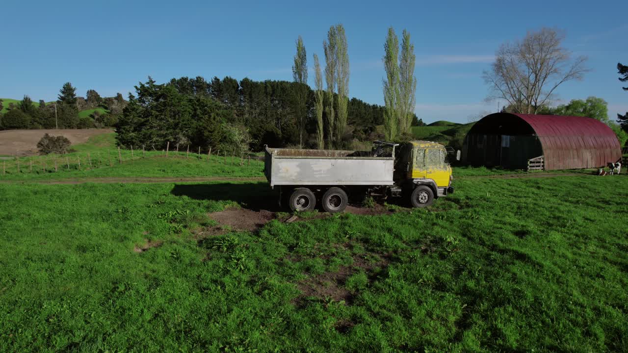 노란색 덤프 트럭이 잔디로 인 농장 에 앉아 있고, 뒷면에는 빨간색 창고와 나무들이 있습니다.