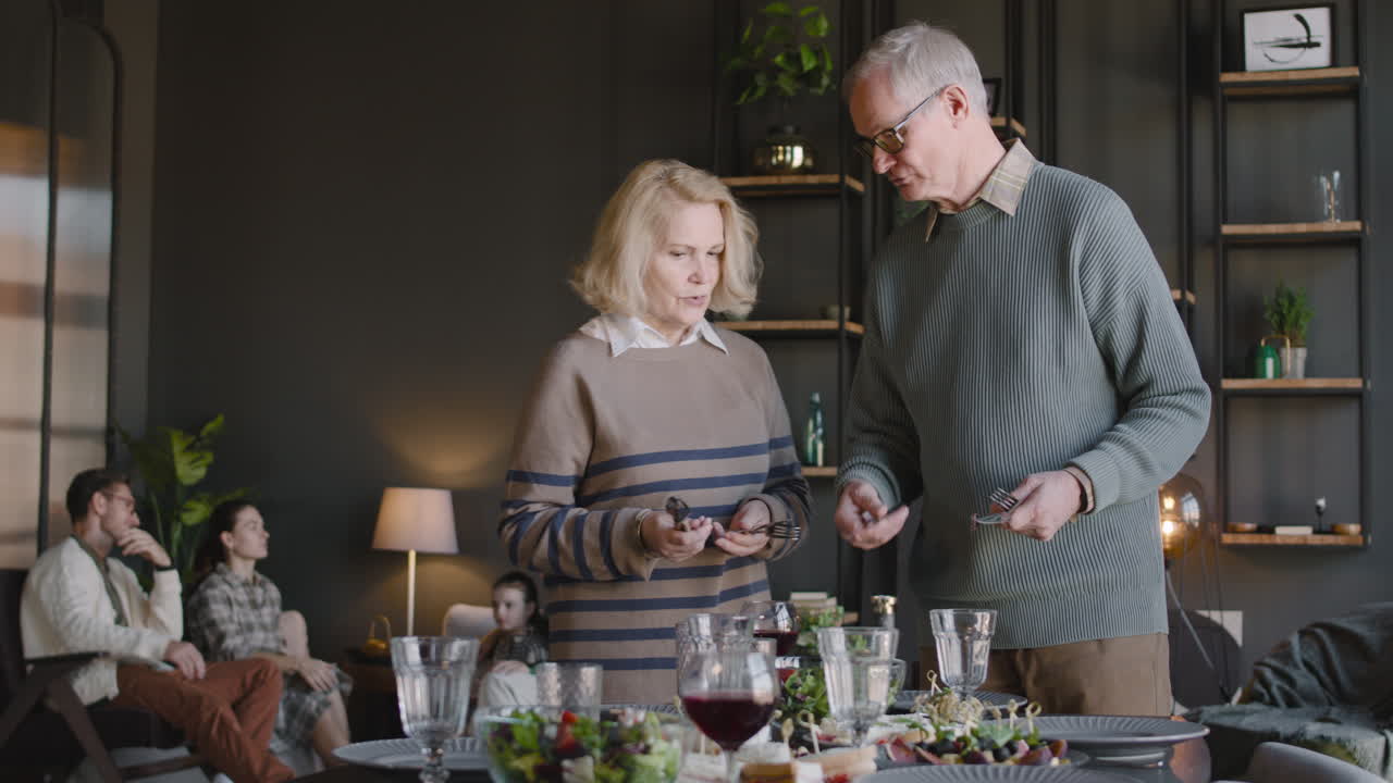 Grandparents Preparing Dining Table, While In The Background Their Family Sitting In Living Room And Talking Together