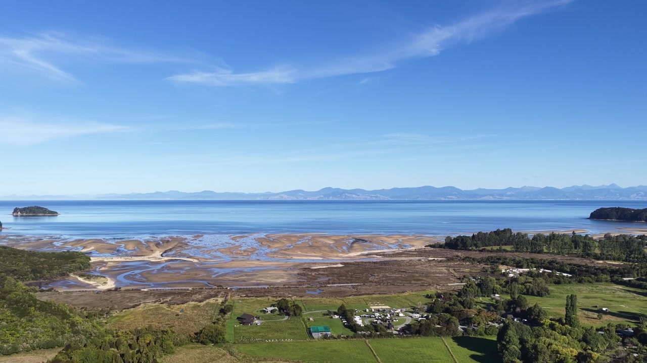 Luftfoto – bred tidevannsestuar med sandbanker, kystby og fjern fjellkjede under blå himmel på Sørøya i New Zealand.