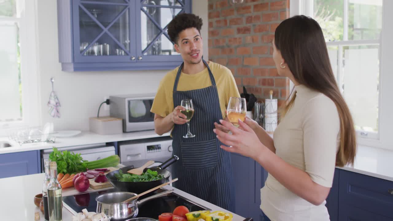 feliz pareja biracial cocinando juntos y bebiendo vino en la cocina