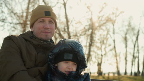 close-up of a father and son sitting closely together on a bench, with the father looking focused, soft sun rays reflect gently behind him, atmosphere, with a blurred background of trees