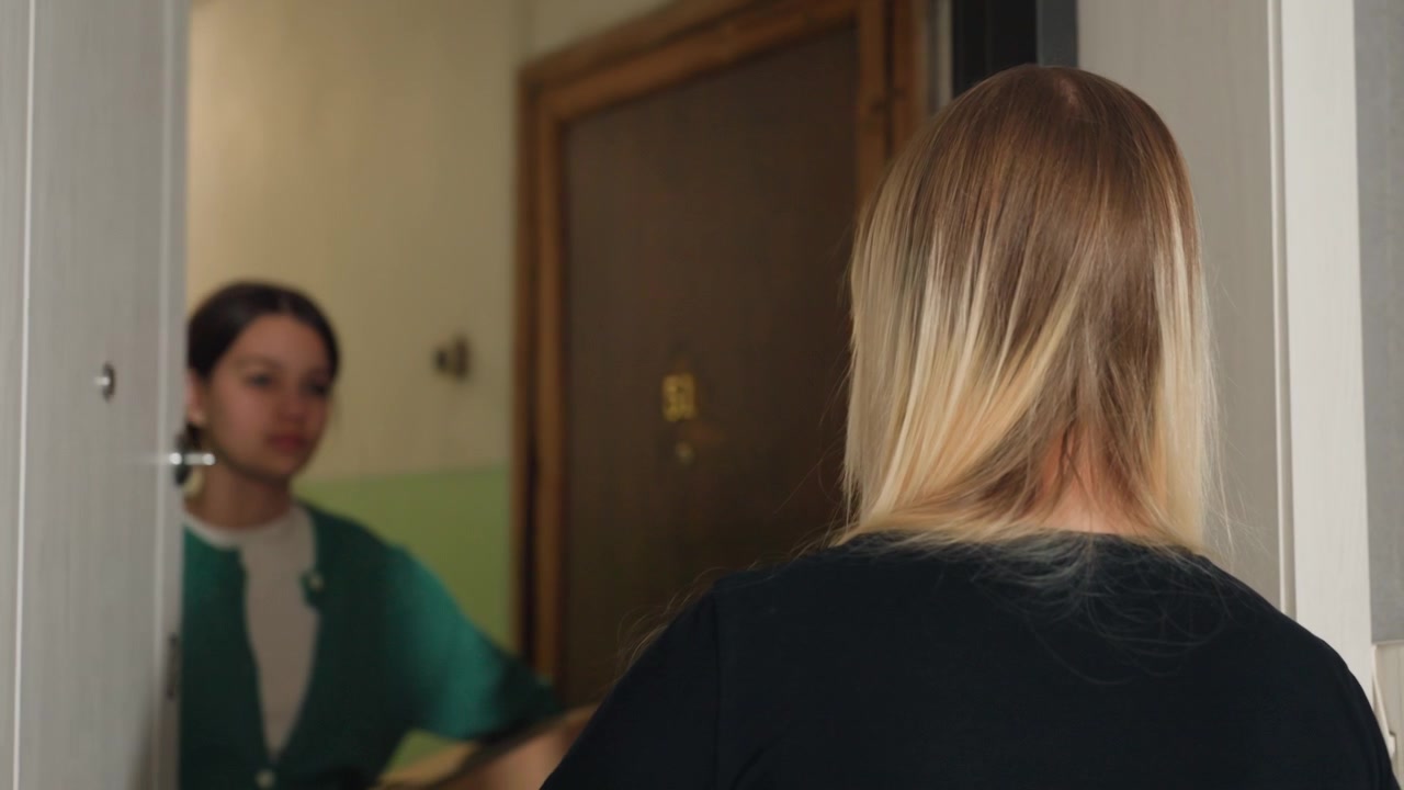 Giovane donna bionda caucasica in piedi nel corridoio in attesa della coinquilina, inquadrature da dietro e di profilo mostrano una tranquilla attesa, illuminazione interna soffusa, griglia a muro a forma di nuvola, messa a fuoco sullo spioncino e sull'ingresso.