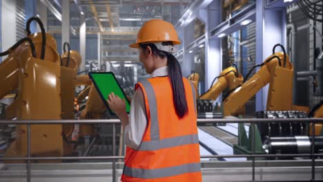 back view of asian female engineer with safety helmet working on a green screen tablet and looking around with industrial robotic arms working