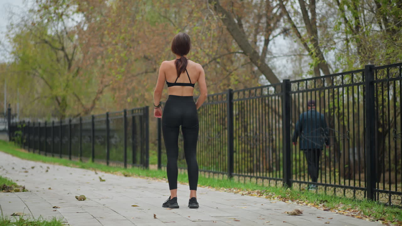 Back view of woman in black fitness wear performing outdoor exercise in front of iron fence with dry trees in background, a man walking outside, creating an active lifestyle scene